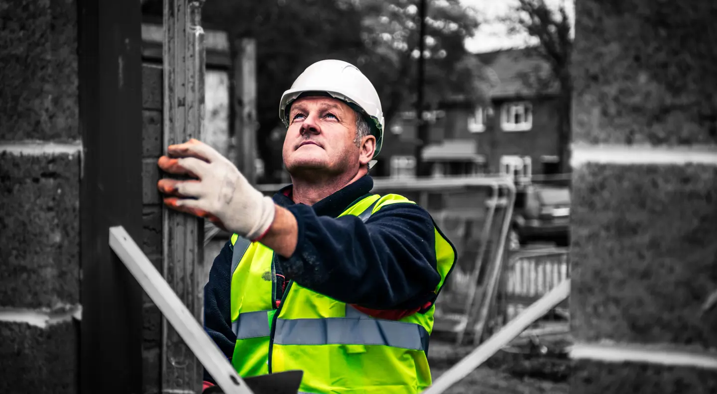 Construction worker wearing a high-visibility vest, hard hat and protective gloves, using measuring equipment at an outdoor building site.
