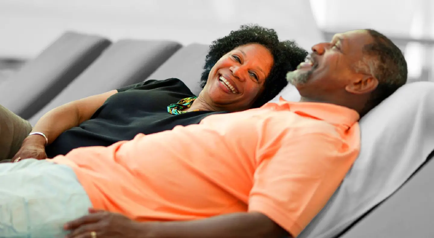 Smiling couple reclining side by side on cushioned lounge chairs, sharing a relaxed, happy moment together.
