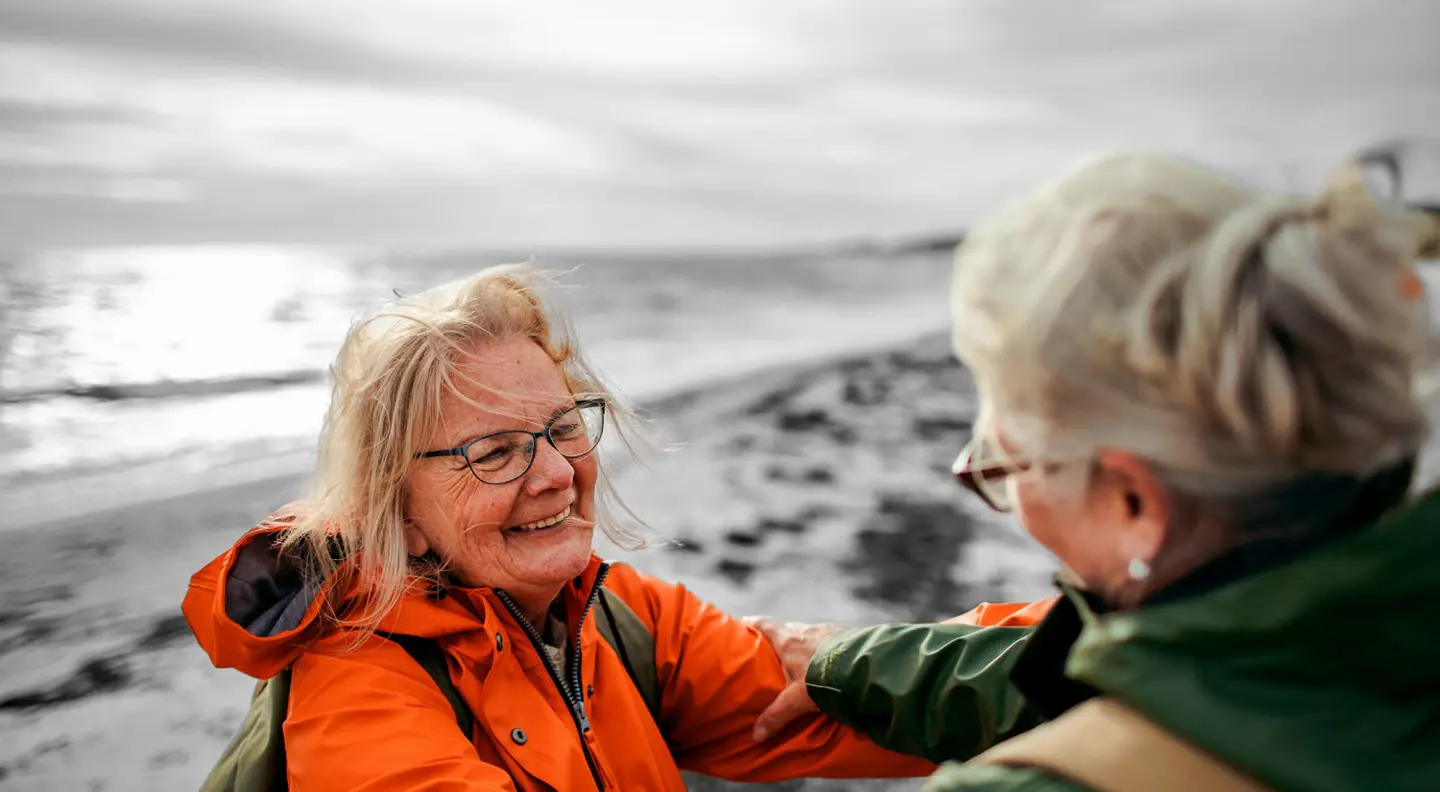 Two people stand on a beach, facing each other and holding onto each other’s arms. One wears an orange jacket and the other a green jacket. The shoreline and ocean stretch out behind them under a cloudy sky, creating a calm coastal atmosphere.
