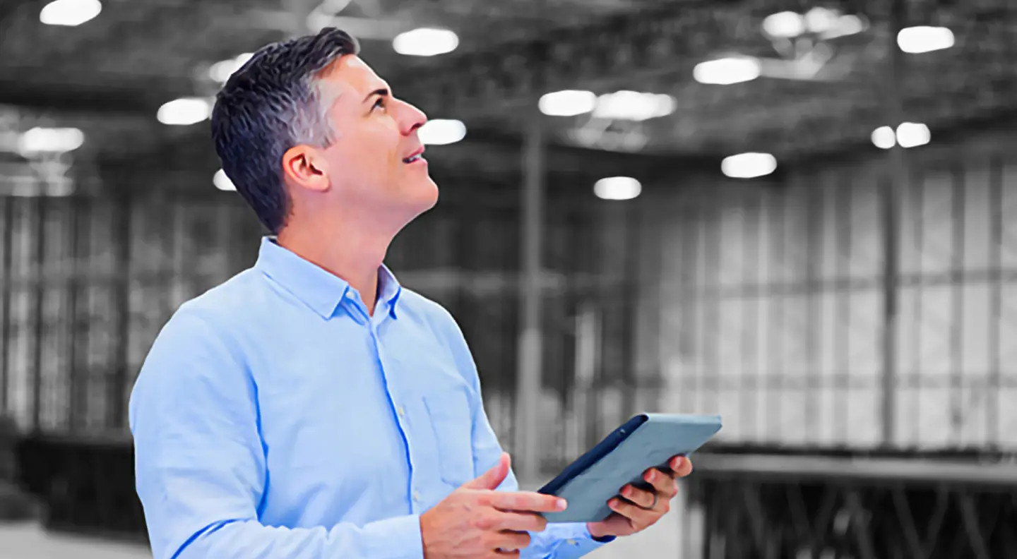 Person holding a tablet while inspecting a large industrial warehouse facility, with warehouse racking and overhead lighting visible in the background.