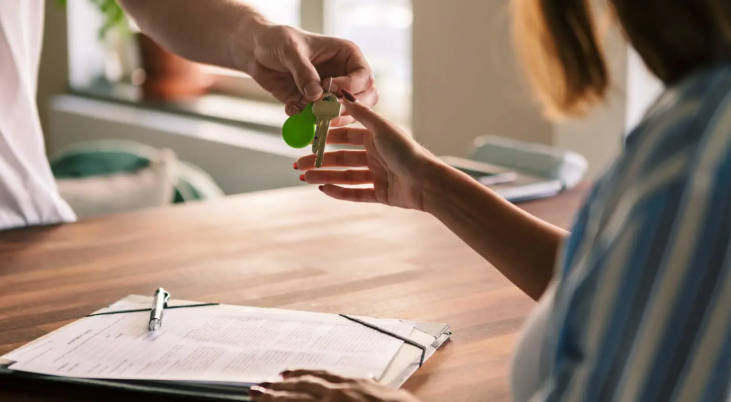 Hands passing a set of keys over a table with paperwork and a pen, in a bright indoor setting.
