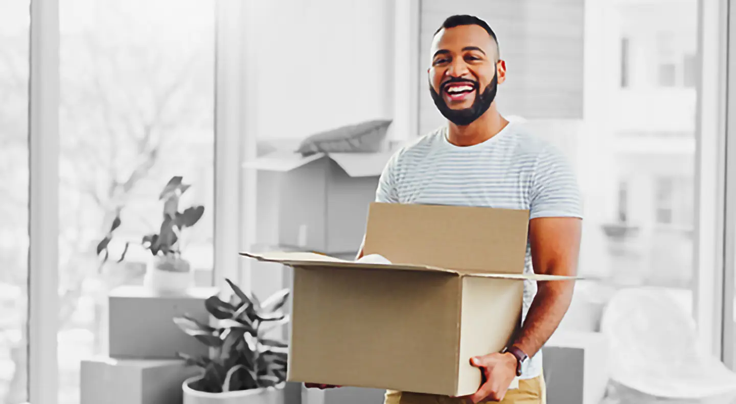 Smiling individual holding two cardboard boxes in a bright living room with white furniture and plants, suggesting a home move.