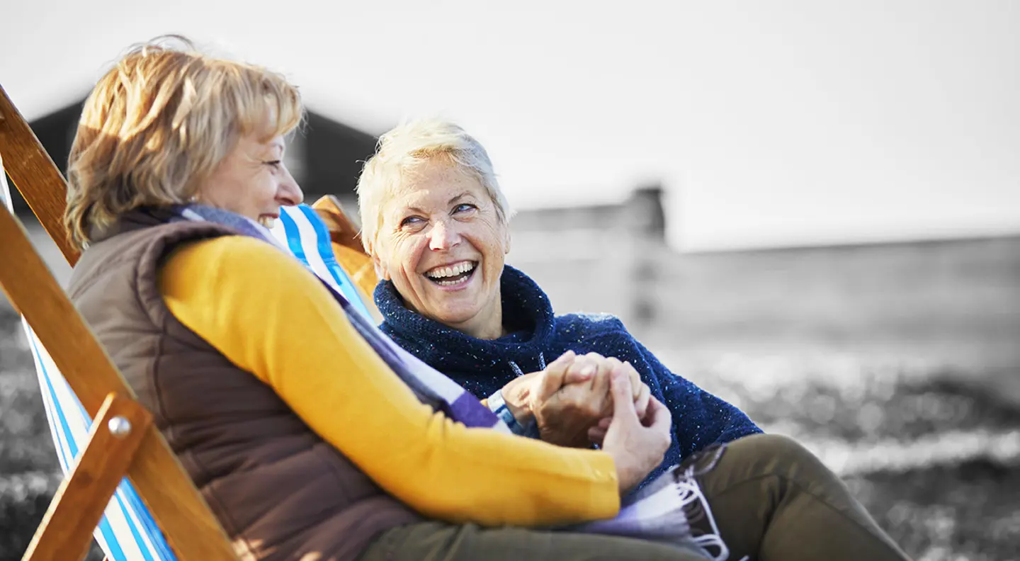 Two people relaxing in deckchairs outdoors, chatting and holding hands in a sunny seaside setting.