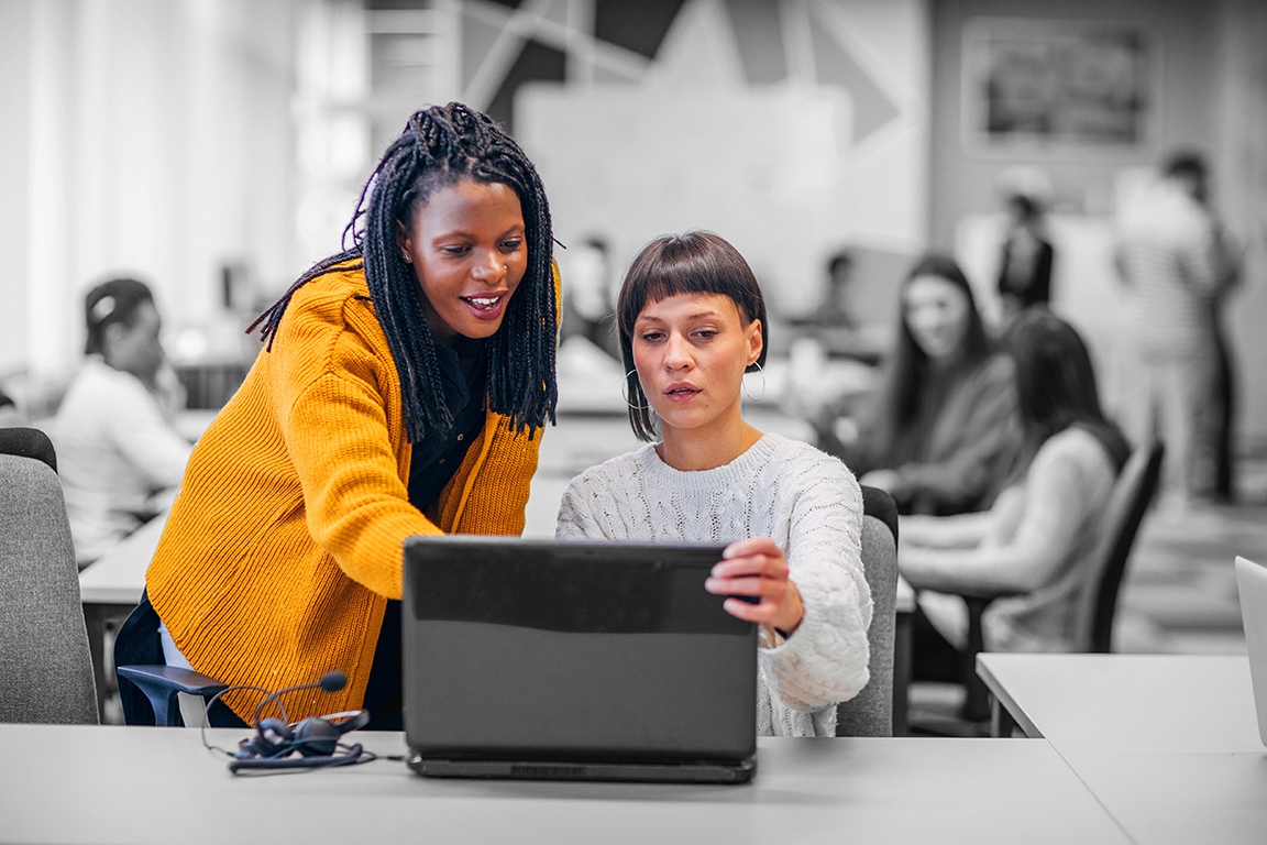 Two coworkers collaborate at a desk, looking at a laptop screen in an open-plan office, while other colleagues work in the blurred background.