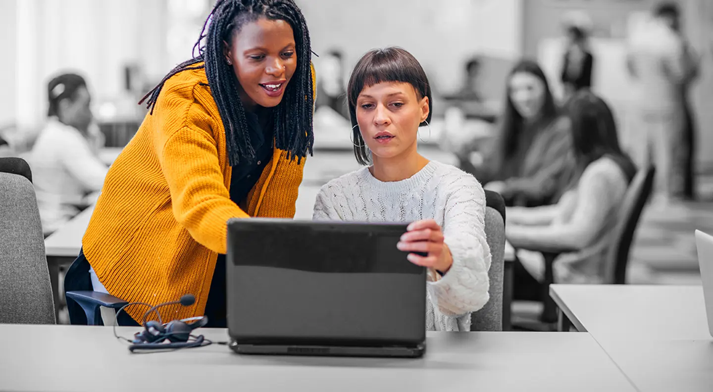 Two coworkers collaborate at a desk, looking at a laptop screen in an open-plan office, while other colleagues work in the blurred background.