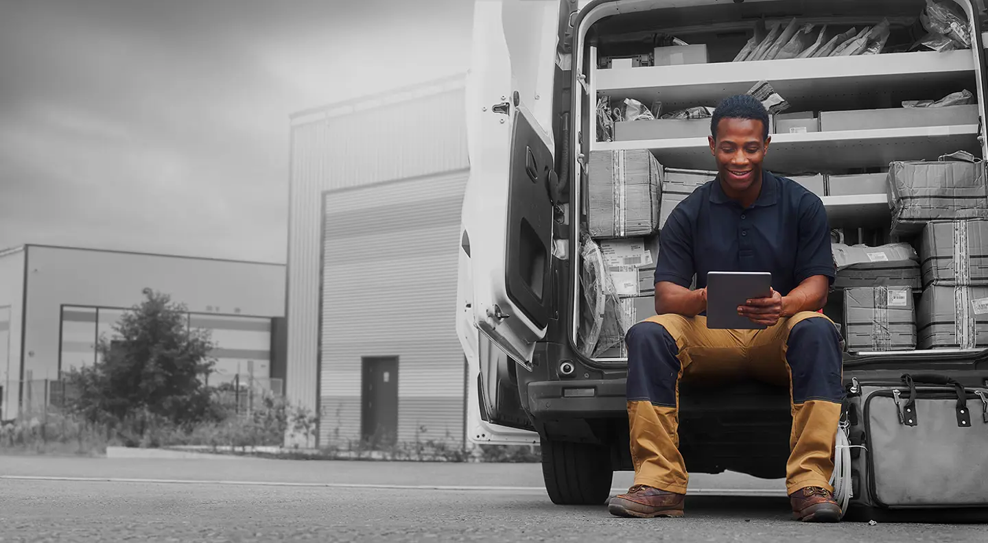 Person sitting on the back of a delivery van using a tablet, with shelves of parcels and packages inside the vehicle. The van is parked outside an industrial warehouse, with most of the background shown in black and white and the person and van highlighted in colour.