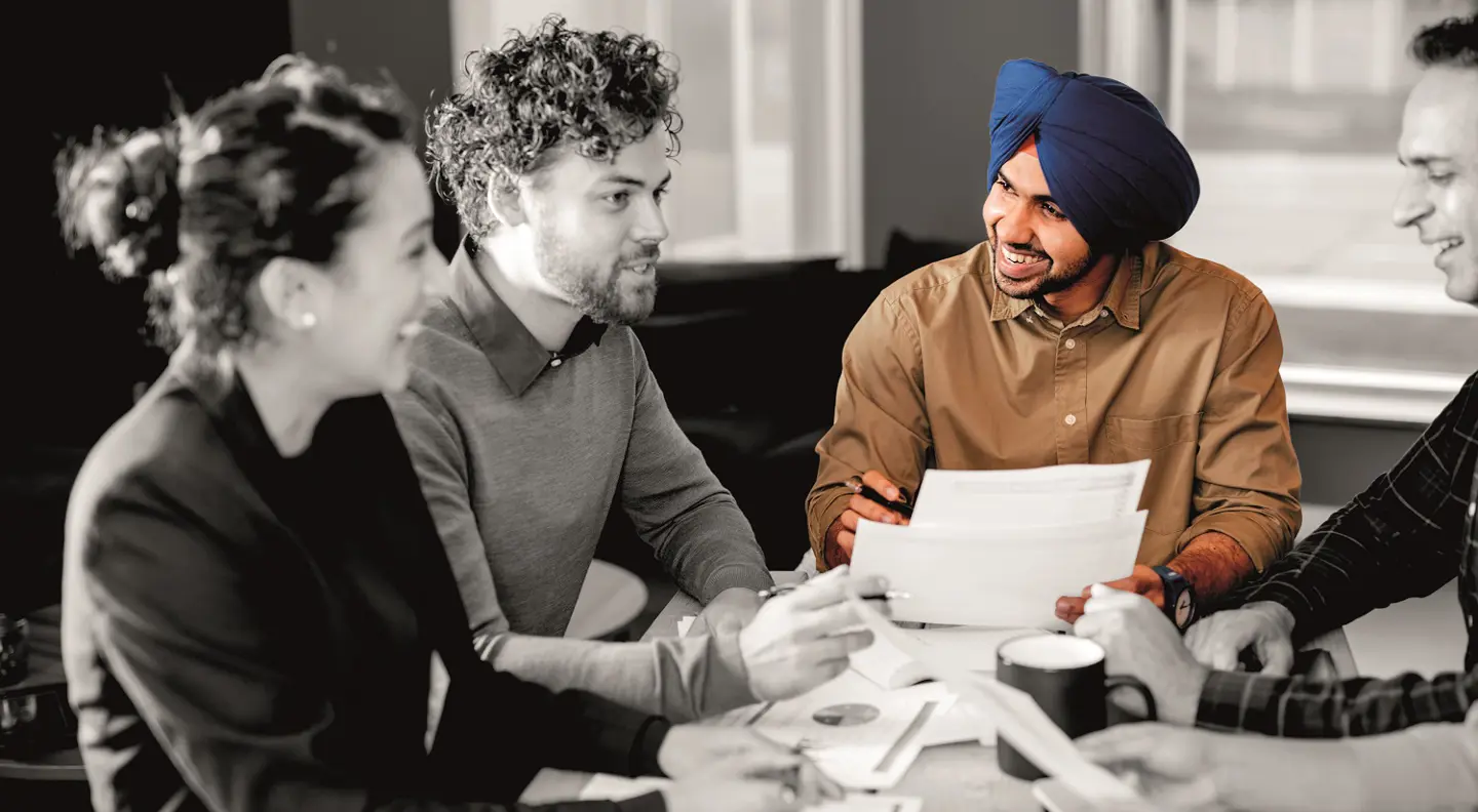Two people sit facing each other across a table in an office environment, one wearing a checked blazer and the other dressed in light-colored clothing, indicating a professional meeting or discussion.