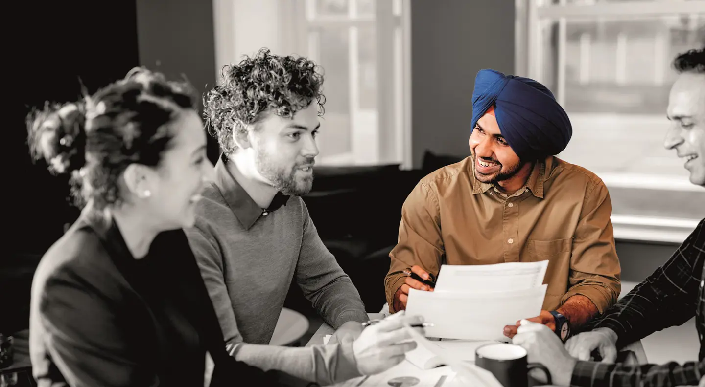 Two people sit facing each other across a table in an office environment, one wearing a checked blazer and the other dressed in light-colored clothing, indicating a professional meeting or discussion.