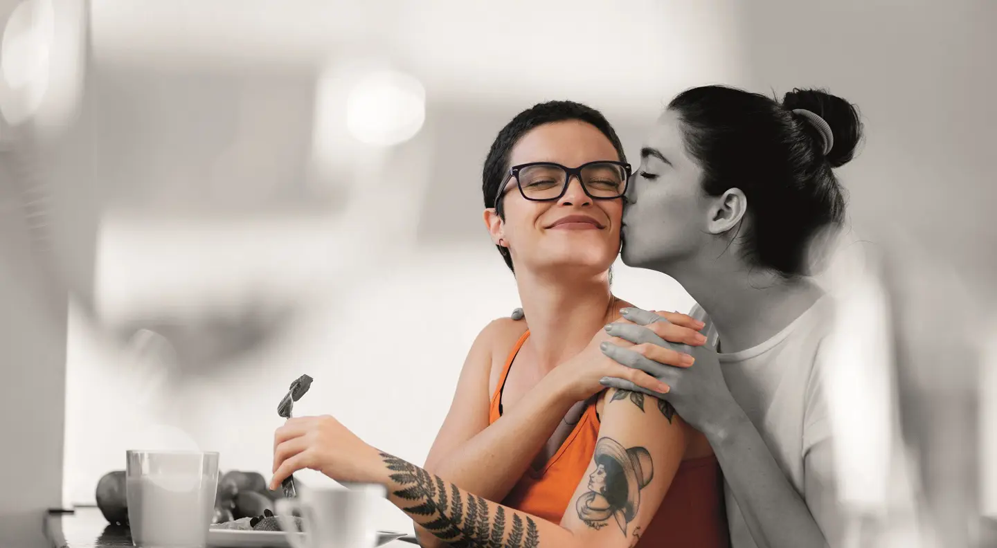 One person sits at a table eating while another stands close behind them, leaning in with their arms wrapped gently around the seated person’s shoulders. The seated person has a visible arm tattoo, and the scene appears warm and affectionate in a softly lit indoor setting.