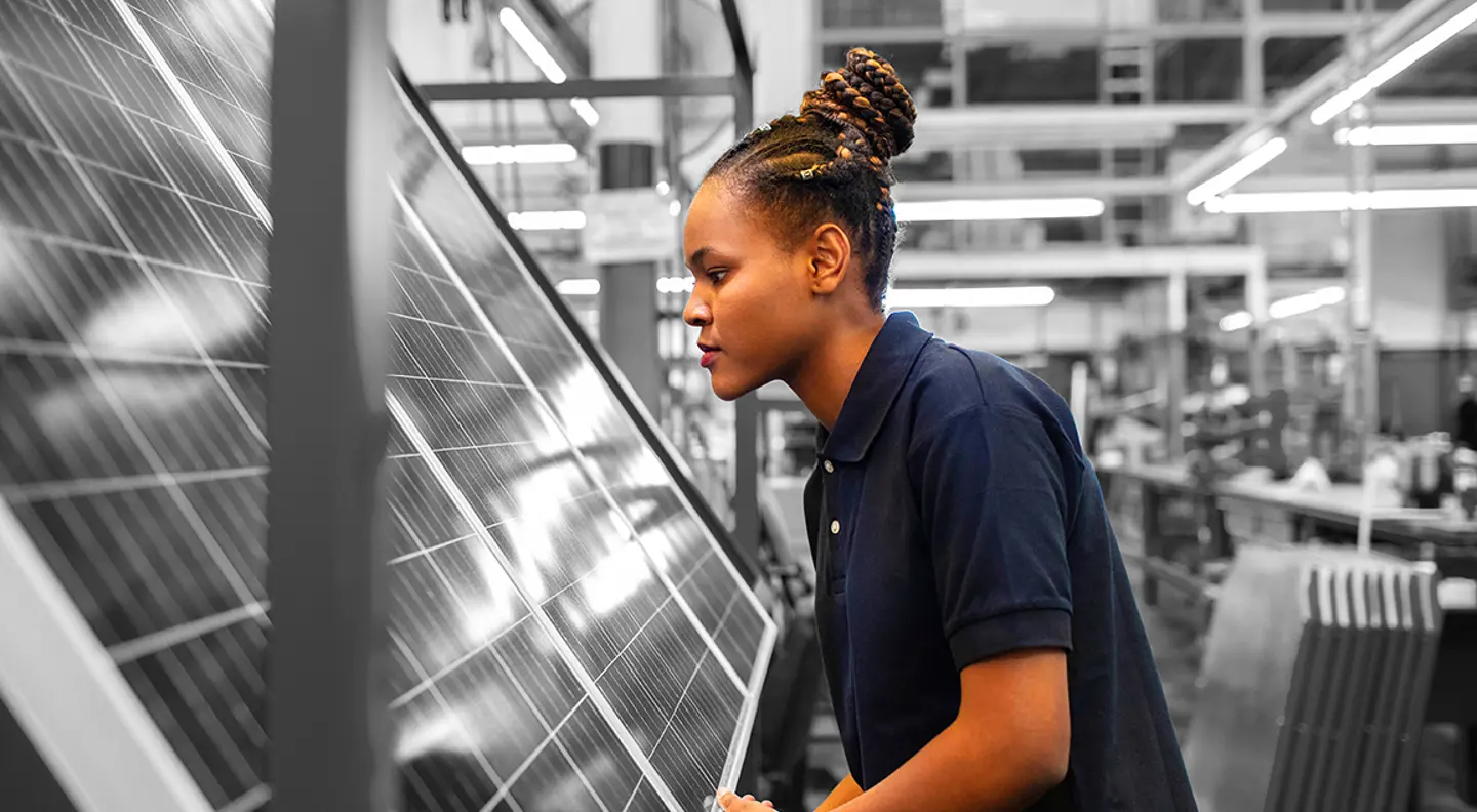 Person inspecting a solar panel in an industrial manufacturing facility, with production equipment and workstations visible in the background.