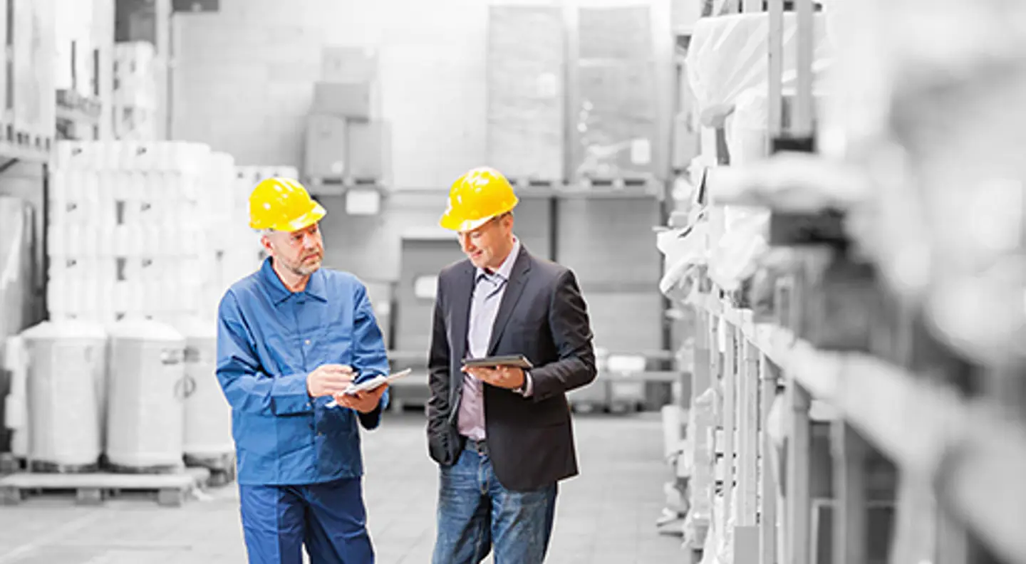 Two people wearing yellow safety helmets walking through an industrial warehouse while reviewing information on a clipboard and tablet. Storage racks and packaged goods line the warehouse, with the workers highlighted in colour against a predominantly monochrome background.