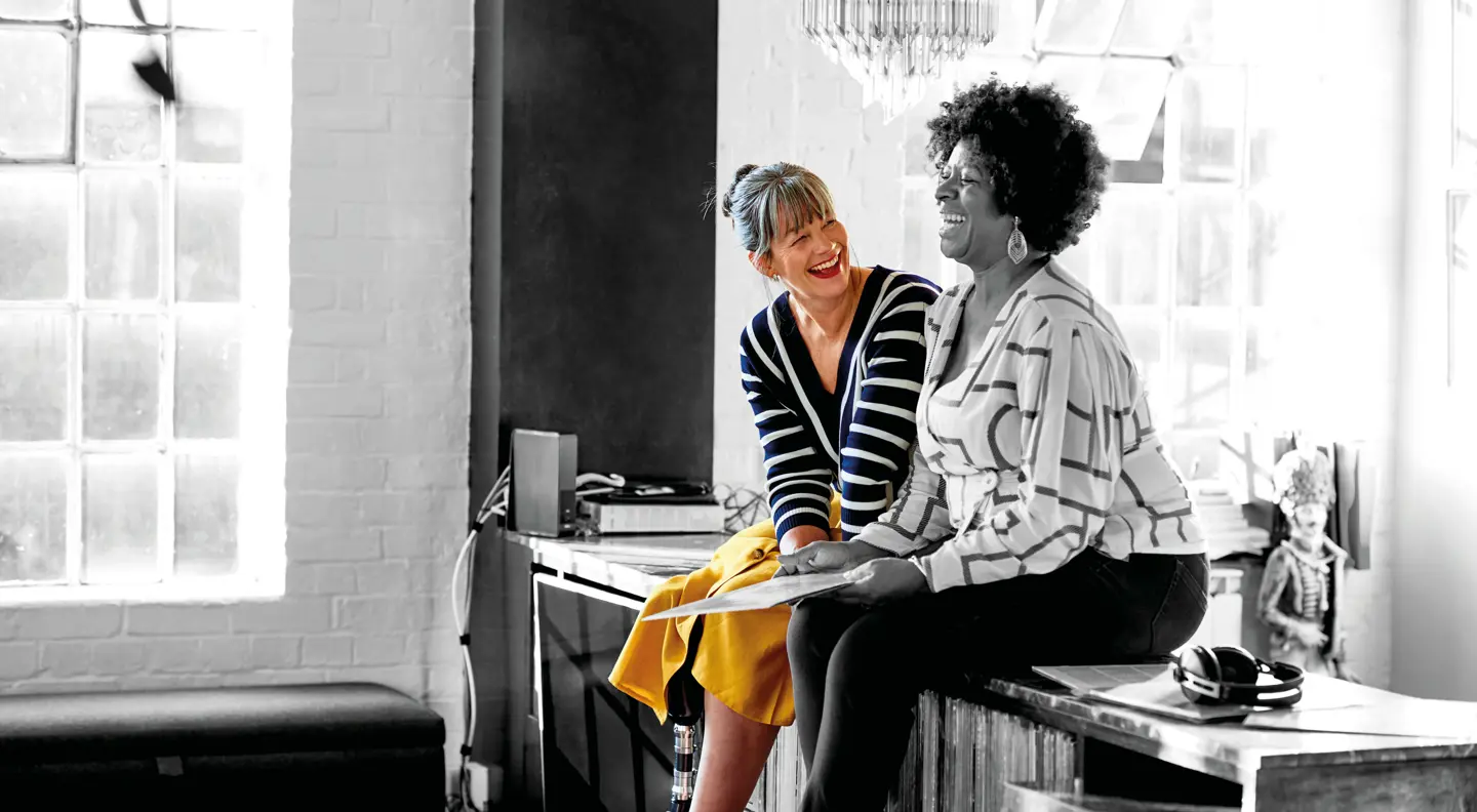 Two people sit on a countertop talking, with one in colour holding papers and the other in grayscale beside them in a bright room.