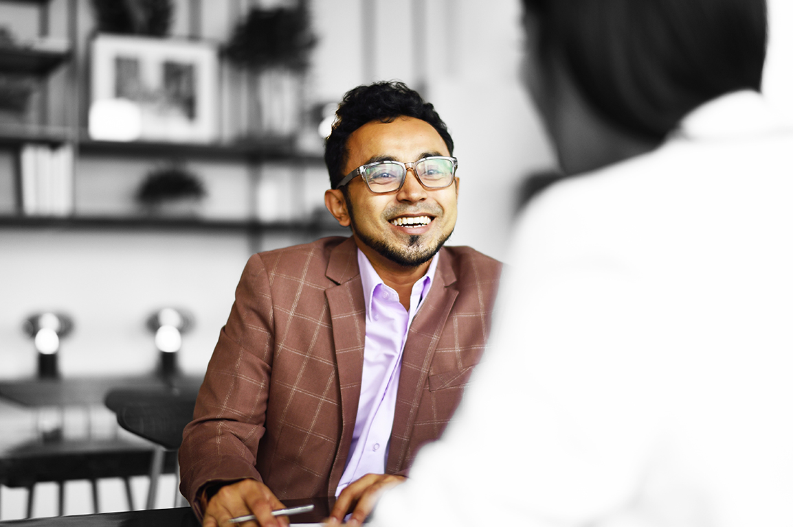Person wearing glasses seated at a table in a professional meeting, engaged in conversation with another individual. A laptop and office furnishings are visible in the background, with the person highlighted in colour against a predominantly monochrome setting.