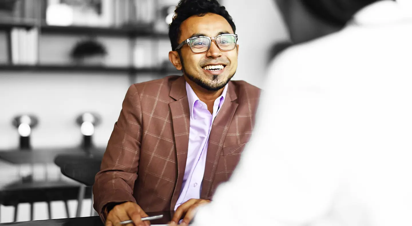 Person wearing glasses seated at a table in a professional meeting, engaged in conversation with another individual. A laptop and office furnishings are visible in the background, with the person highlighted in colour against a predominantly monochrome setting.